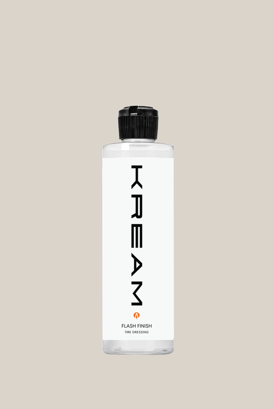 A sleek spray bottle of Kream Auto’s Flash Finish Tire Dressing, featuring a modern, minimalist white label with the brand name "KREAM" in bold, vertical black letters. The clear bottle shows a white liquid inside, and the product is labeled as “Flash Finish Tire Dressing” with a small orange logo and the tagline “Clean smarter, not harder.” The bottle has a black squeeze nozzle, set against a soft gray background.